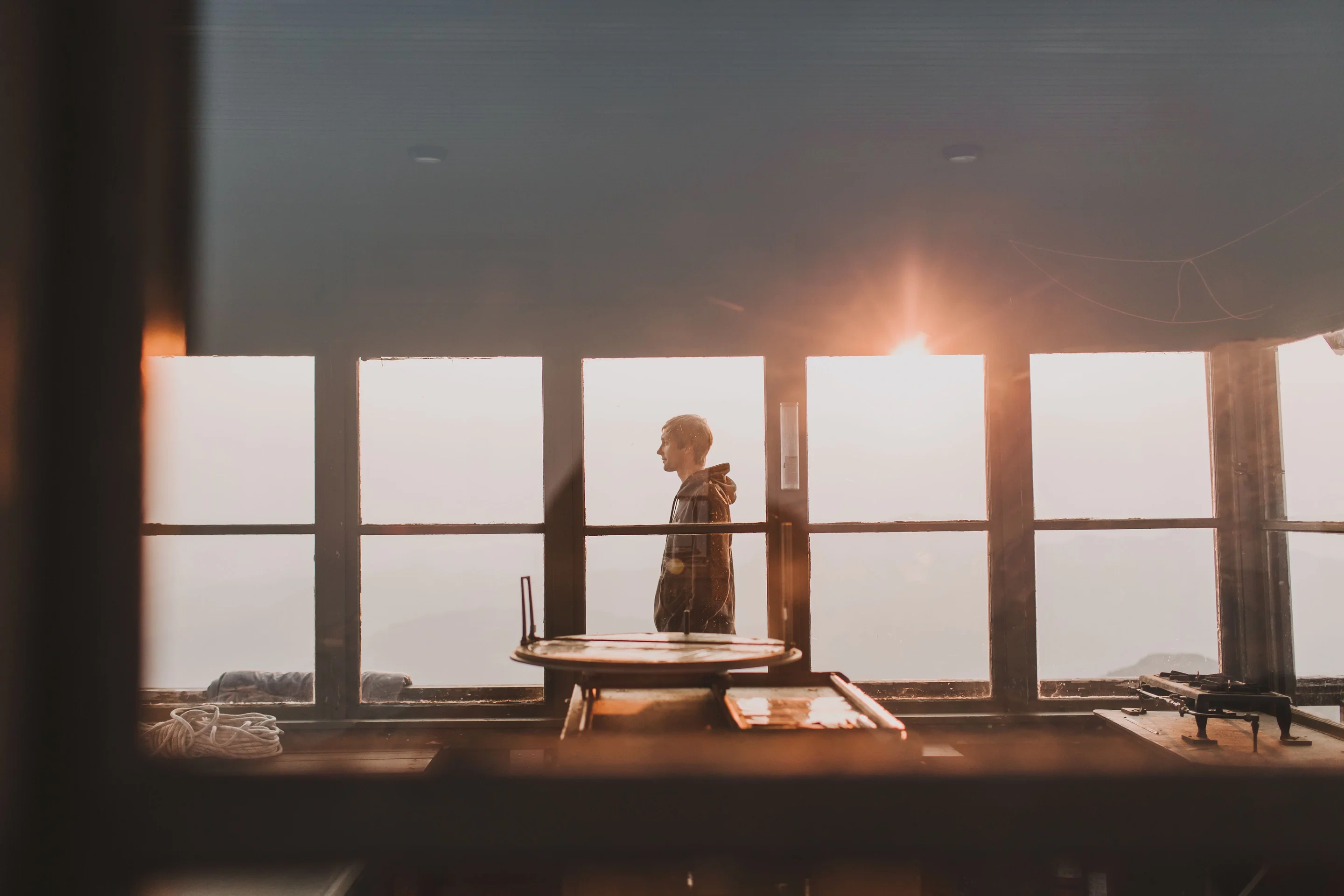 A man standing in a mountain fire-lookout at sunset, looking out over the hills — a quiet moment of freedom and peace.